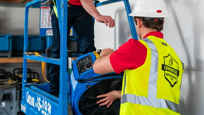 Two Rainbow Restoration service professionals lifting an industrial-strength fan onto a lift in a warehouse.