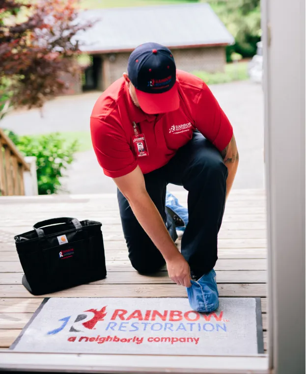A Rainbow Restoration service professional putting on shoe coverings at a customer's front door next to a branded company mat and briefcase.