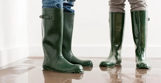 A couple in Lochbuie, CO, wears rubber boots and stands in a flooded living room surrounded by water-damaged carpet before calling a water damage restoration company.