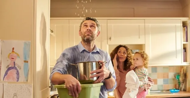 A man stands in a kitchen in their home in Anderson, SC, and catches water falling from the ceiling with a large pot. A woman holding a child is on the phone calling a water damage restoration company.