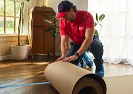 A Rainbow Restoration service professional unrolling a large sheet of heavy-duty paper on a customer's wood floor to protect it during restoration work.