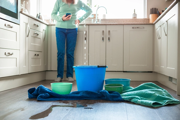Woman stands in the kitchen holding a phone to call a restoration company during a water damage emergency as water falls from the ceiling.