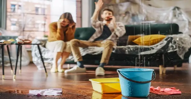  A man and woman sit on a sofa to call a water damage restoration company in Thornton, CO, as water falls from the ceiling and collects in a container on the floor.