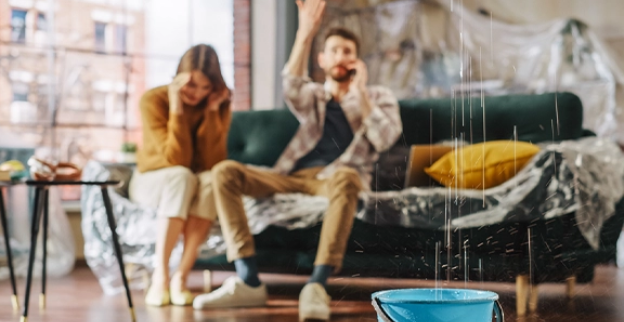  A man and woman sit on a sofa to call a water damage restoration company in Thornton, CO, as water falls from the ceiling and collects in a container on the floor.