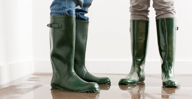 Two people wearing rubber boots and stands in a flooded living room surrounded by water-damaged carpet.