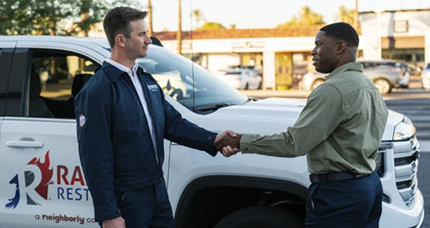 A Rainbow job manager stands beside a Rainbow Restoration pickup truck, greeting an office manager.