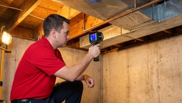 A restoration expert in a red polo and navy pants uses an infrared camera to distinguish between ductwork condensation and a structural pipe leak in a Canton basement.