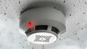 A smoke detector with a red warning light is mounted on a speckled ceiling, representing the hidden smoke damage that can remain after a home fire.