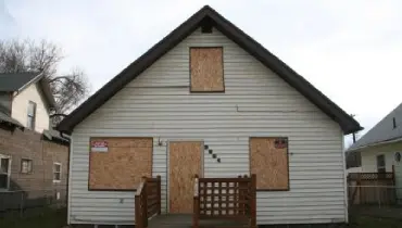 A white two-story house with boarded-up windows and doors, showing plywood coverings over all openings.