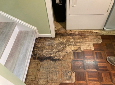 A partially removed section of flooring near a laundry area reveals dark water damage and mold beneath old parquet tiles. The photo shows discolored subflooring with visible staining next to a staircase and a dryer, indicating moisture intrusion and deterioration.