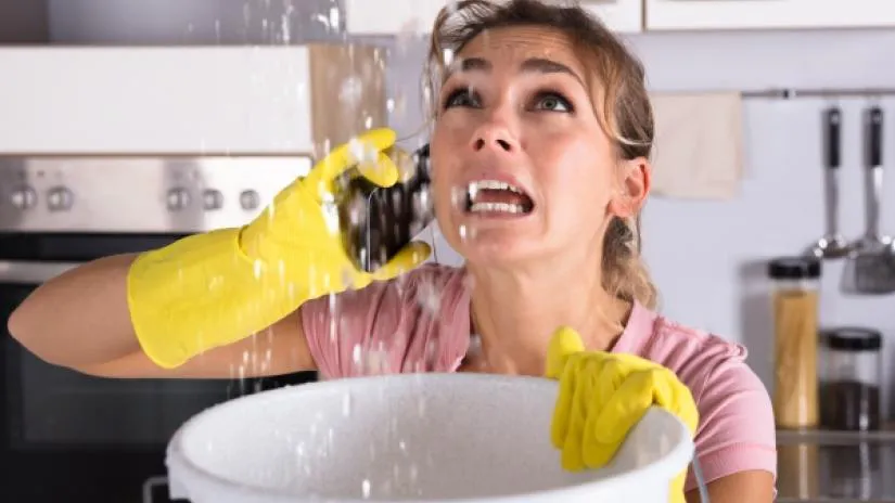 Woman wearing yellow rubber gloves, holding a phone and looking distressed as she catches water in a bucket that is leaking from the ceiling.