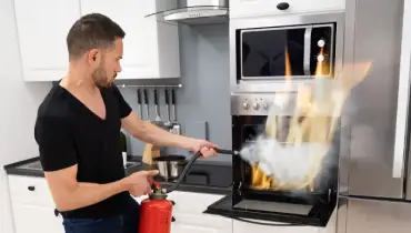 A man uses a fire extinguisher to put out flames inside a kitchen oven, illustrating the risk of kitchen fires that can lead to smoke and structural damage.
