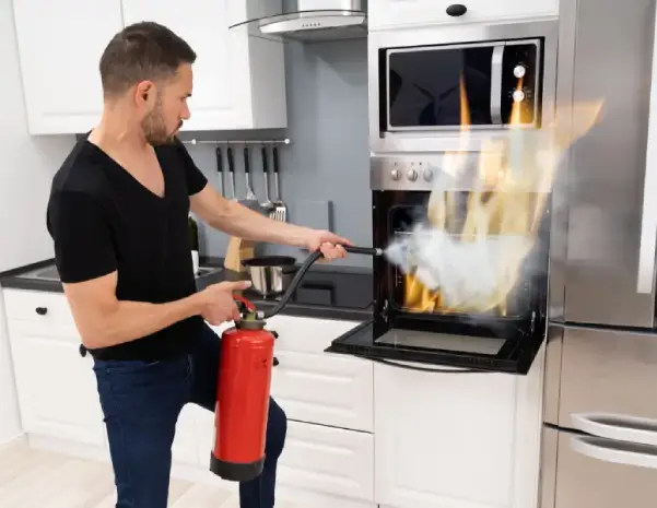 A man uses a fire extinguisher to put out flames inside a kitchen oven, illustrating the risk of kitchen fires that can lead to smoke and structural damage.