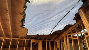 A storm-damaged roof is temporarily covered with a protective tarp above an exposed residential interior.