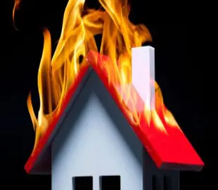 A small model house with a red roof is shown engulfed in bright orange flames against a dark black background, symbolizing a house fire.