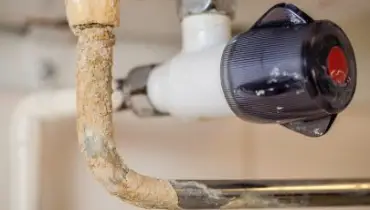 A close-up view of a corroded and mineral-encrusted pipe beneath a sink, showing signs of water damage and buildup near a plastic shut-off valve with a red indicator.