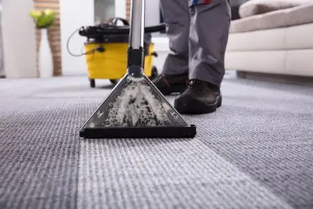 A professional technician uses a commercial carpet cleaning machine to deep-clean a gray carpet in a living room. The cleaning head is visibly extracting dirt and moisture, with a yellow cleaning unit and hose in the background.