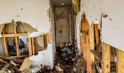 A heavily damaged interior of a home shows torn drywall, exposed wooden framing, and debris scattered across the floor. Signs of severe water damage and contamination are visible throughout, highlighting the aftermath of a flood or extensive leak.
