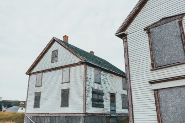 Two old white wooden houses with weathered exteriors are shown with all windows securely boarded up using plywood. 