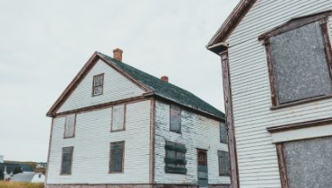 Two old white wooden houses with weathered exteriors are shown with all windows securely boarded up using plywood.
