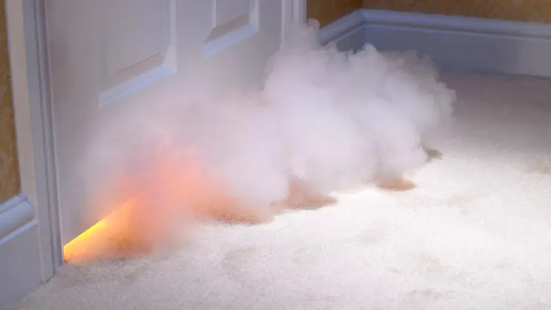 Smoke billows from the crack between a door and a carpeted floor, with fire in the room on the other side of the door.